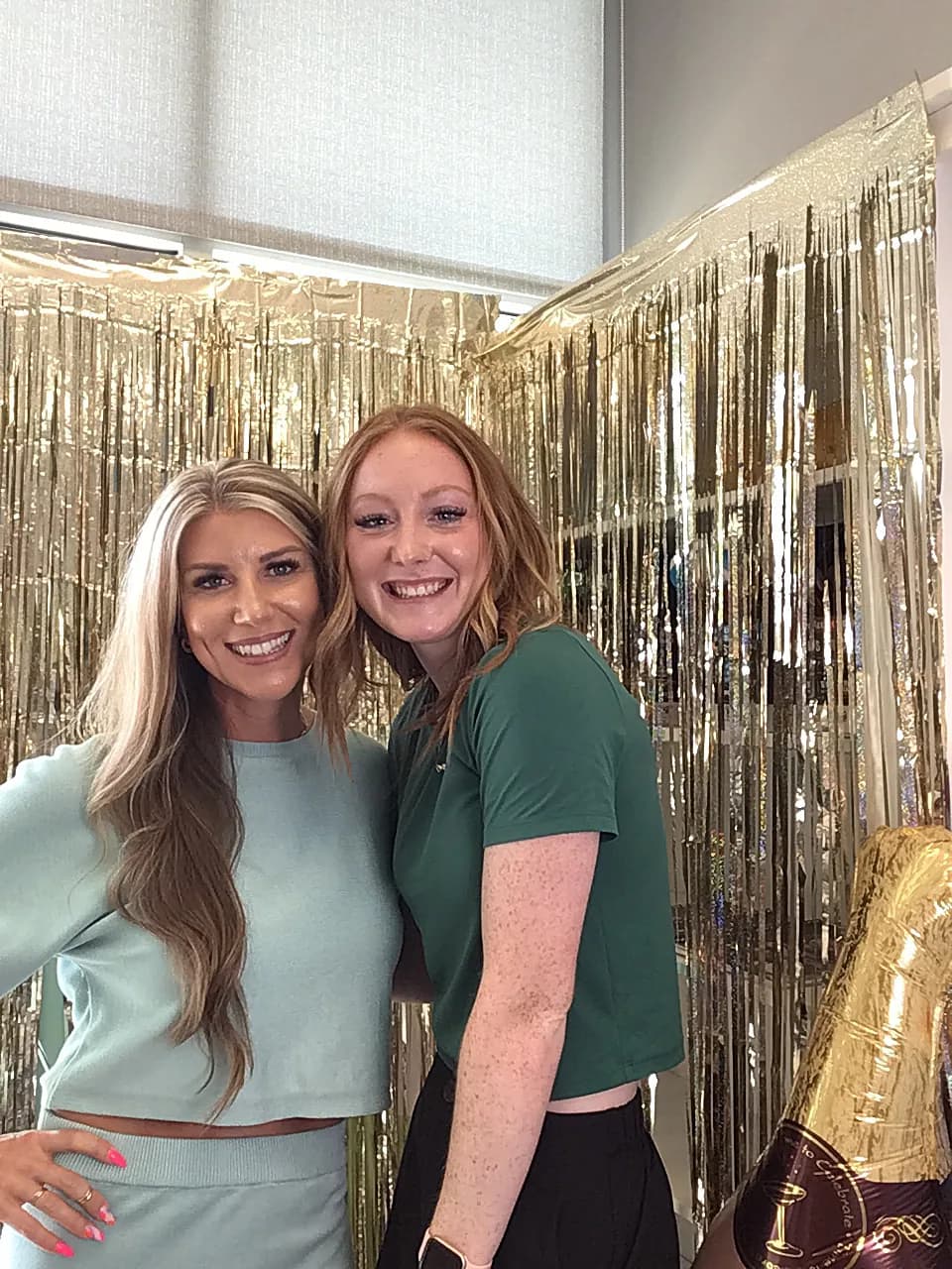 Two team members smiling in front of the gold fringe photo wall